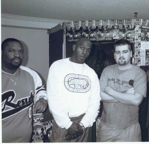 Black-and-white 1990s photograph of a young DJ Wrekk with two friends in a room papered with rap flyers and posters.