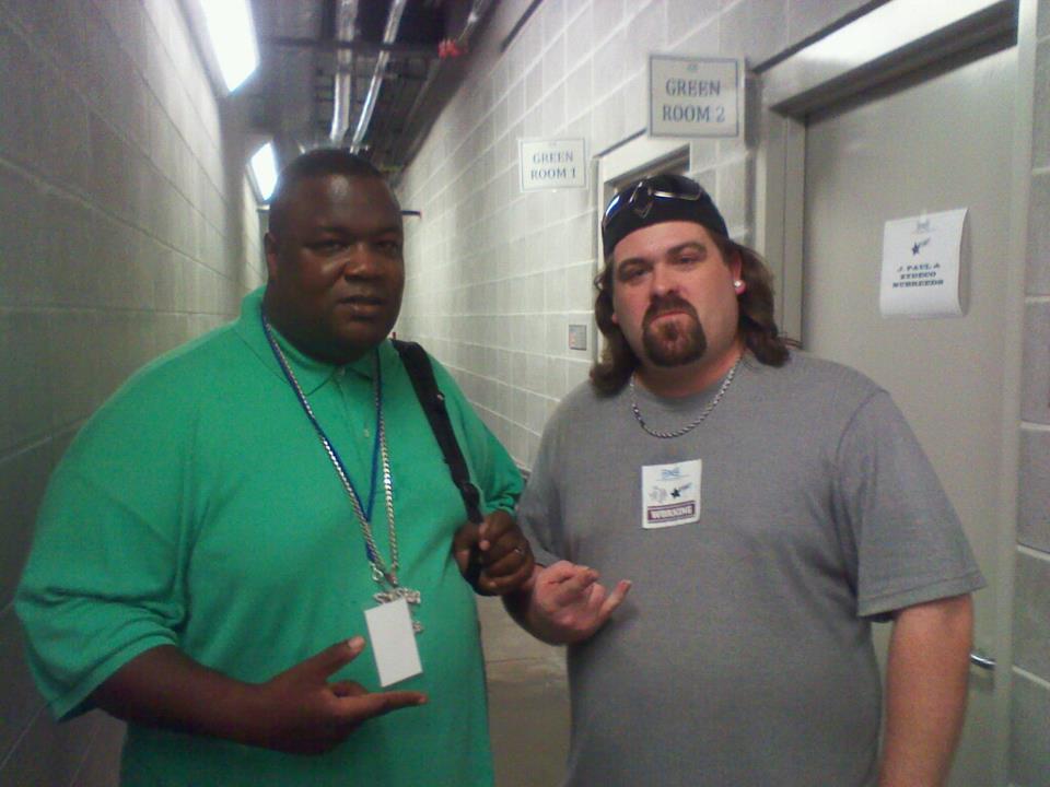 DJ Wrekk and a friend backstage in a cinder-block hallway between two green rooms.