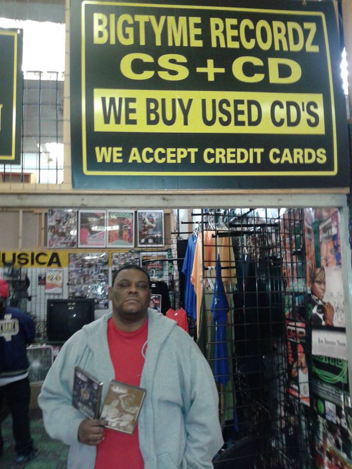 A man holding CDs beneath a yellow Bigtyme Recordz 'CS + CD' sign inside a Houston record shop.