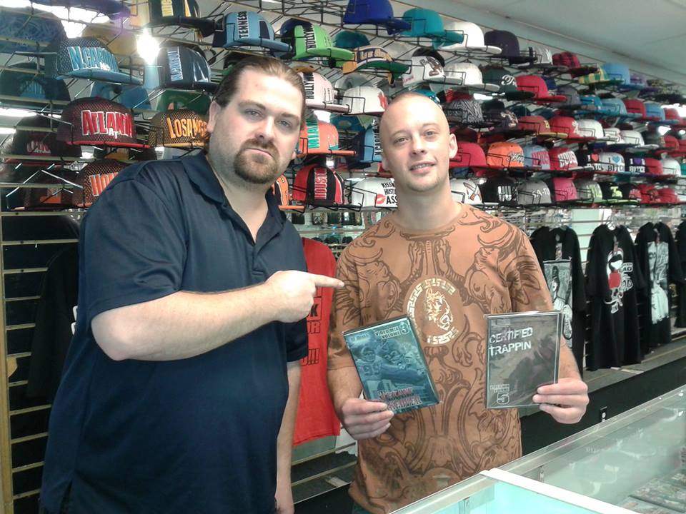 DJ Wrekk pointing to a friend who holds up two mixtape DVDs, in front of a wall of fitted caps at a Houston shop.