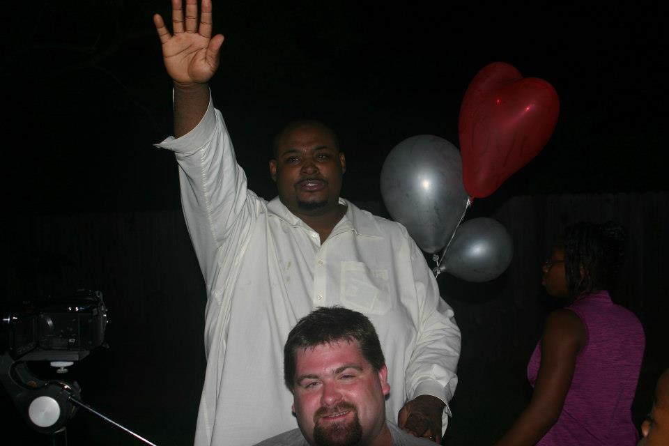 A friend in a white shirt waves to the camera over DJ Wrekk's shoulder at a nighttime backyard party with balloons.