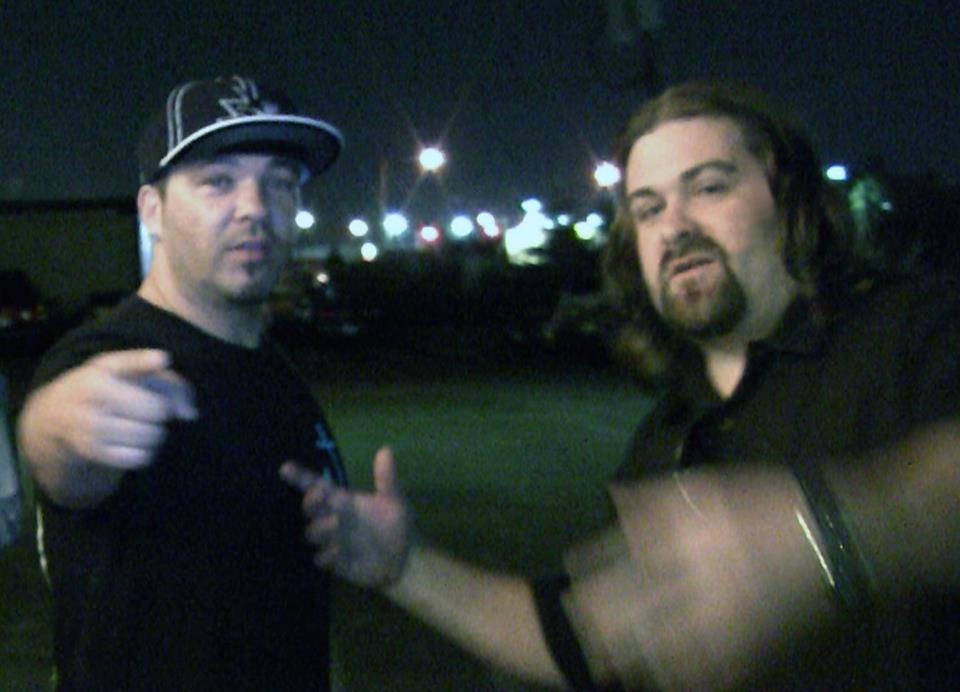 DJ Wrekk and a friend mugging for a camcorder in a parking lot at night, lot lights flaring behind them.