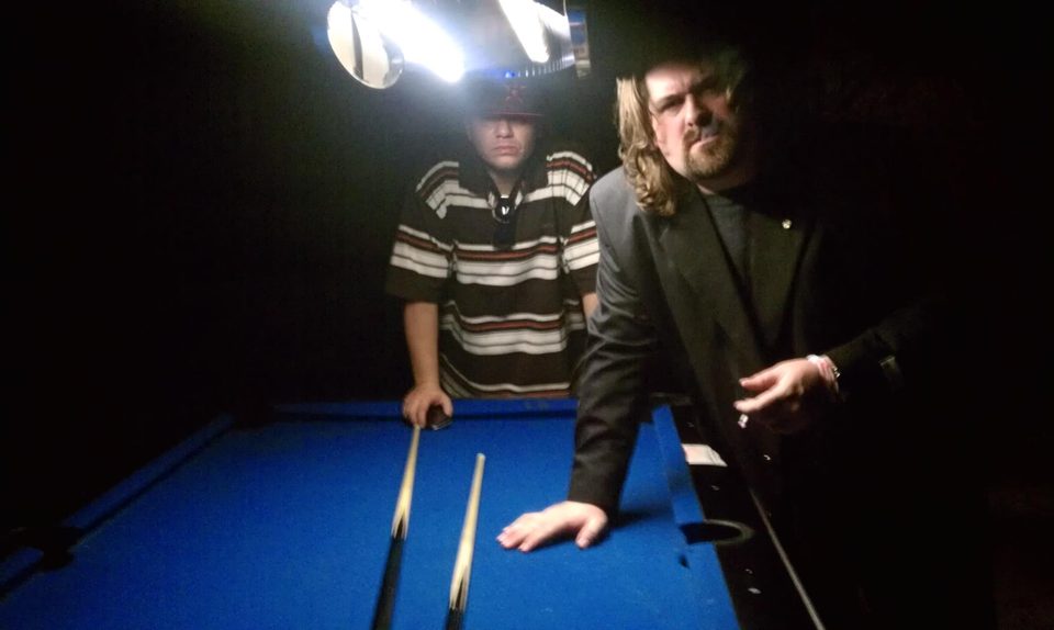DJ Wrekk and Jose 'Block of Rock' DeLeon leaning over a blue pool table under a hanging lamp, cue sticks laid out between them.